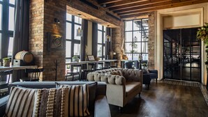 A spacious room with a high ceiling, exposed wooden beams, large windows, a fireplace, and a seating area with a sofa and chairs.