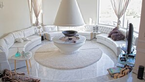 A modern living room with a central circular fireplace, white sofas, and a glass coffee table. - Villa Kokalis Resort (Hersonissos)