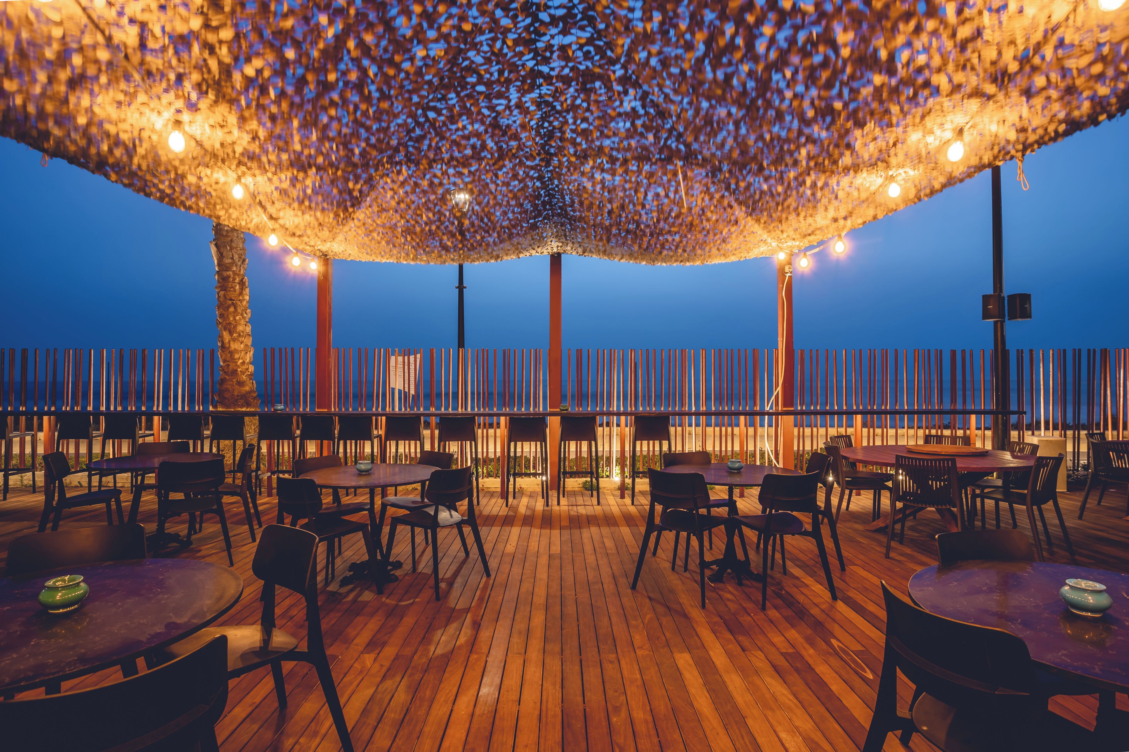 An outdoor dining area with wooden tables and chairs, a pergola with string lights, and a view of the sea.