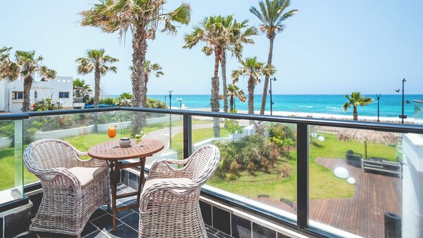 A balcony with wicker chairs, a table, and a glass railing overlooking a beach with palm trees and a clear sky. - Sea life Nahariya BY Jacob Hotels (Nahariya)