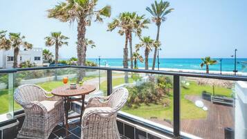 Ein Balkon mit Korbstühlen und einem Tisch, mit Blick auf einen Strand mit Palmen und einem klaren Himmel.