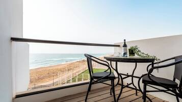 Un balcón con vista a la playa, dos sillas, una mesa con una botella y vasos, y una planta.