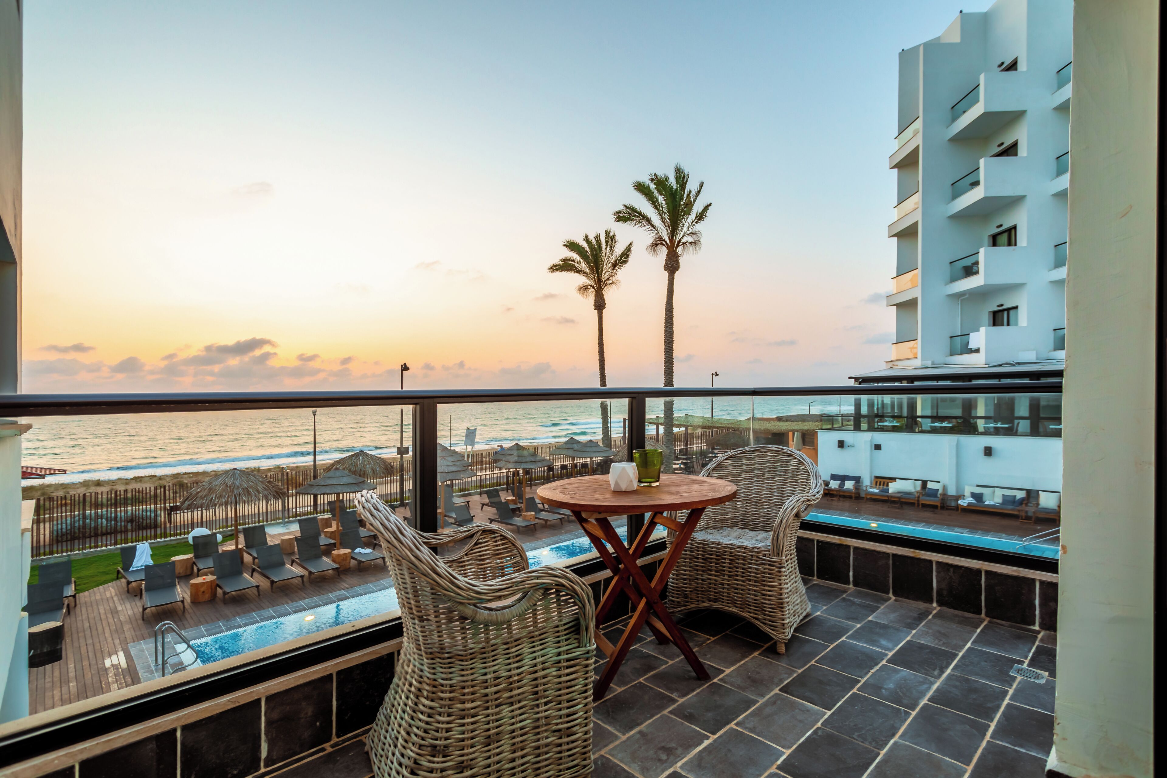 Un balcon avec une piscine, des chaises longues et une vue sur la plage et l’océan au coucher du soleil.