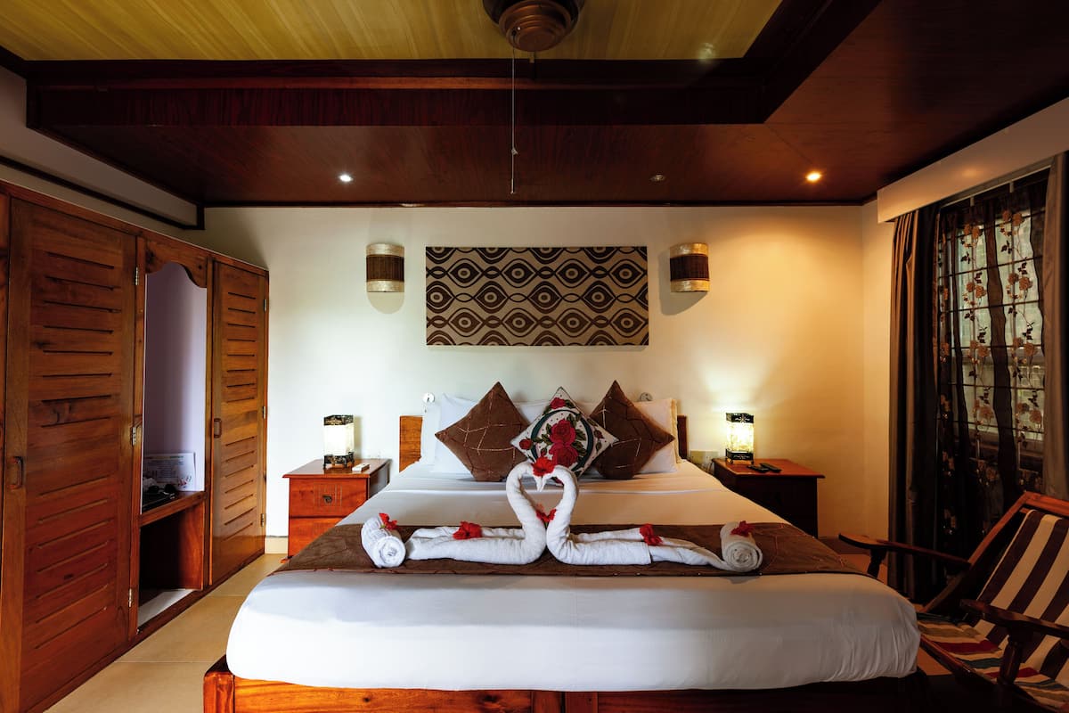 Quarto de hotel com uma cama grande, armário de madeira, duas mesas de cabeceira com luminárias, um objeto decorativo na parede e uma janela com cortinas.