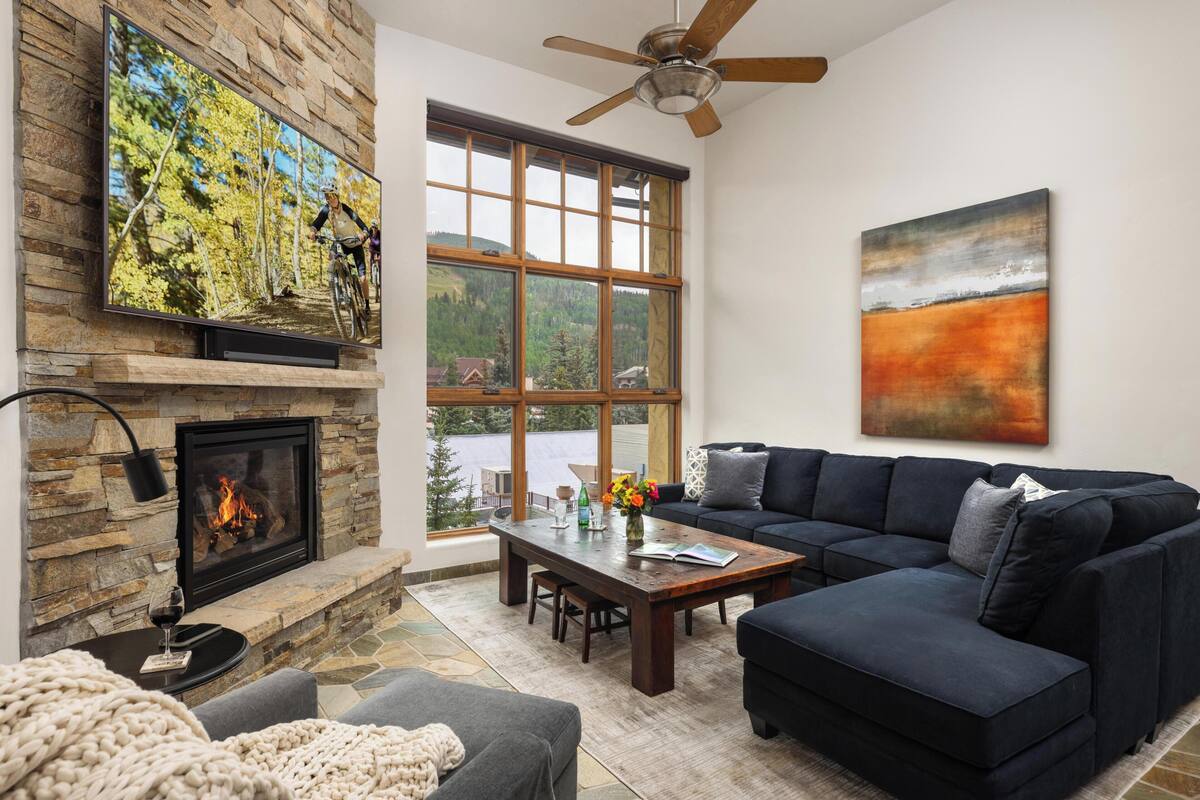 A living room with a stone fireplace, a large TV mounted on the wall, a modern sofa, and a large window with a view of a lake.