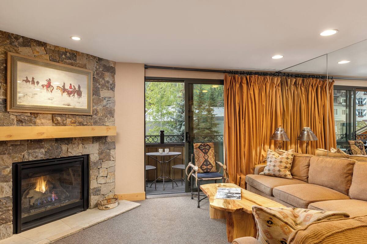 A living room with a stone fireplace, a sofa, a coffee table, and a balcony with a table and chairs.