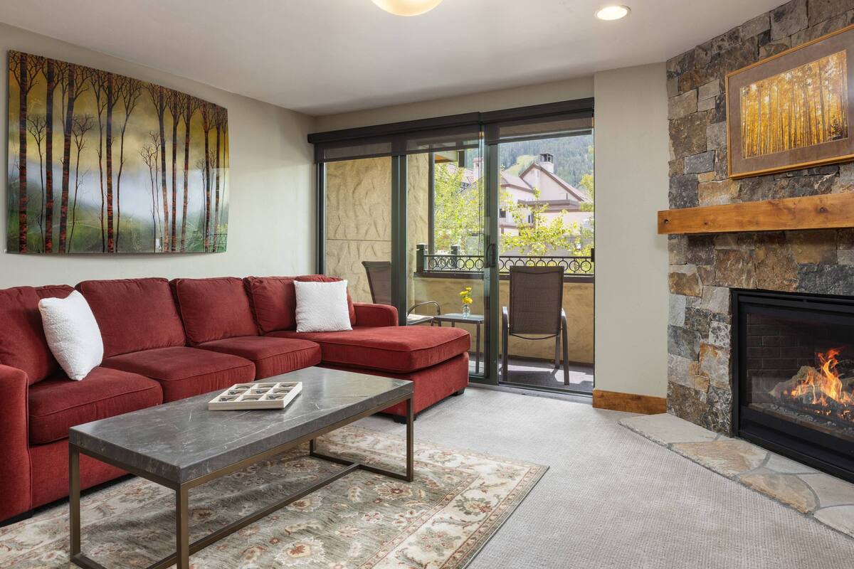 A living room with a red sofa, a coffee table, a fireplace, and a large window with a view of buildings and trees.