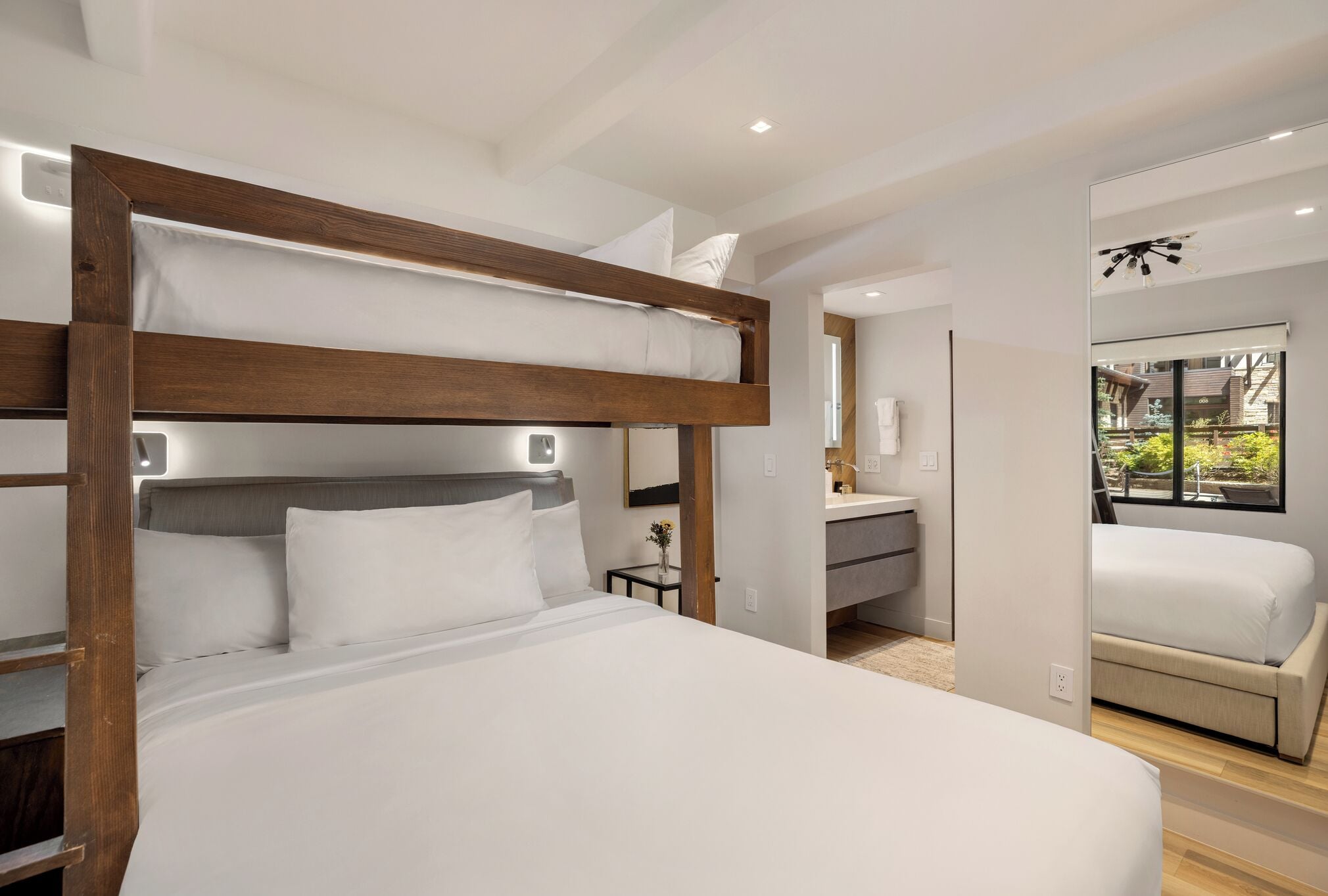A modern hotel room with a bunk bed, a small bedside table with a vase, and a view of the garden through a window.