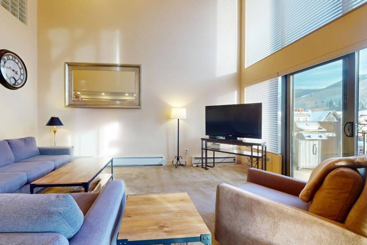 A modern living room with a sofa, coffee table, TV, and large windows offering a view of the outdoors.