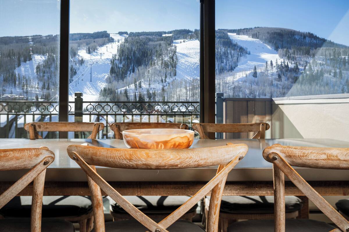 A dining area with wooden chairs and a table, overlooking a snowy mountain landscape through large windows.
