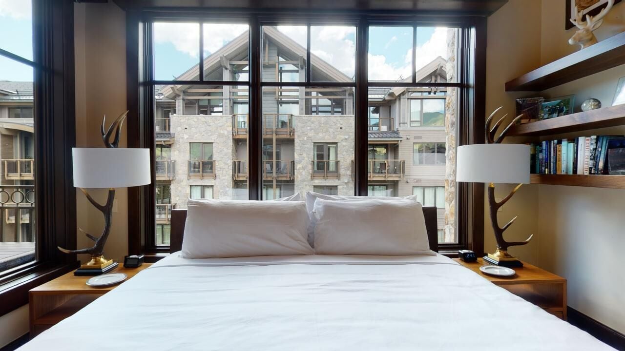 Hotelkamer met een groot bed, twee bedlampen, een nachtkastje en uitzicht op de buitenkant van het gebouw door grote ramen.