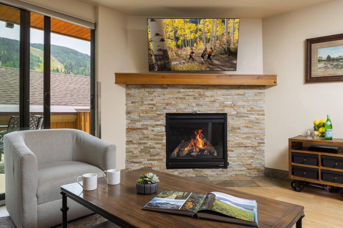 A cozy living room with a stone fireplace, a comfortable sofa, a coffee table with magazines, and a large window offering a scenic view.