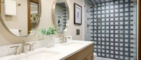 Une salle de bain avec deux lavabos, un grand miroir, une plante en pot et une douche dont le mur est recouvert de carreaux à motifs.