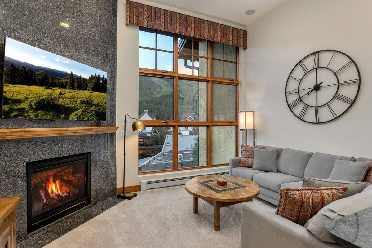 A living room with a fireplace, a large window, a sofa, and a wall clock.