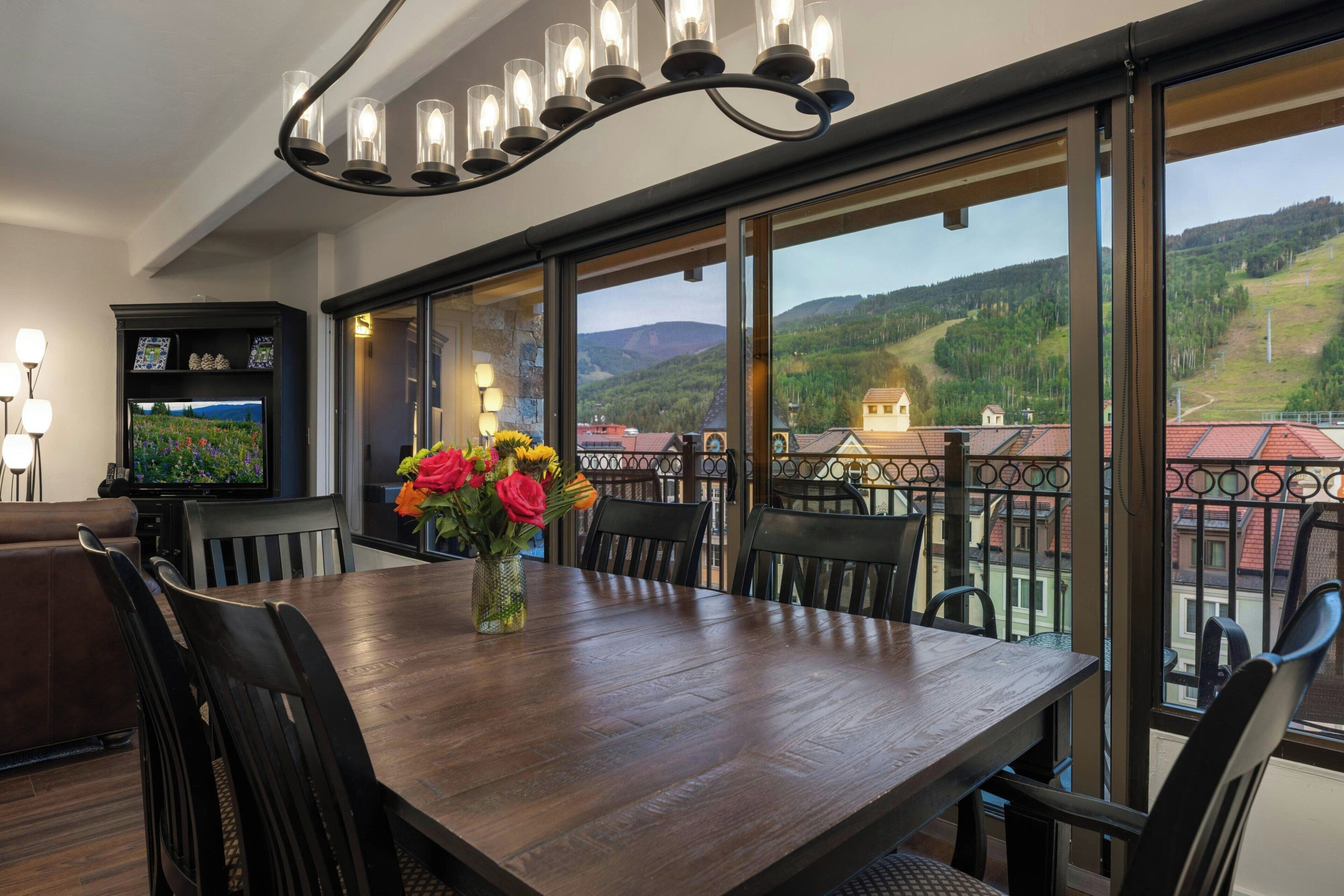 A dining area with a large table, chairs, a vase of flowers, a television, and a balcony with a view of the countryside.