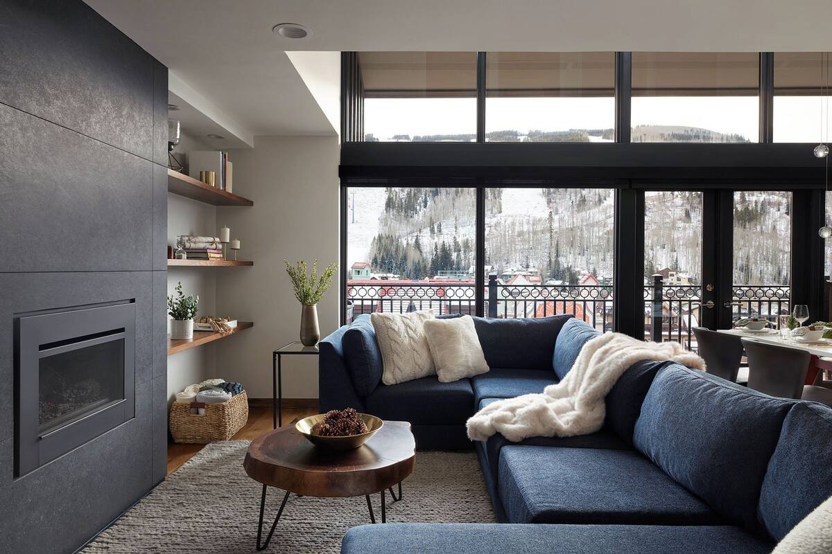 A modern living room with a dark gray wall, a built-in fireplace, a blue sofa, and a large window overlooking a snowy landscape.