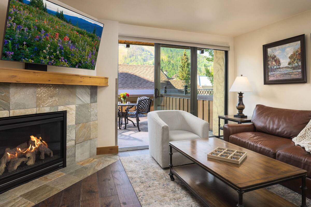 A living room with a fireplace, a television mounted on the wall, a sofa, a coffee table, and a view of a balcony with outdoor seating.