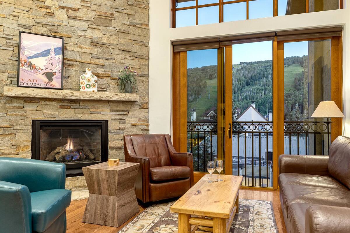 A cozy living room with a stone fireplace, leather sofas, and a large sliding glass door leading to a balcony with a scenic view.