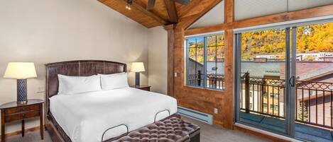 A bedroom with a large bed, two bedside tables with lamps, a ceiling fan, and a view of a balcony with autumn foliage.