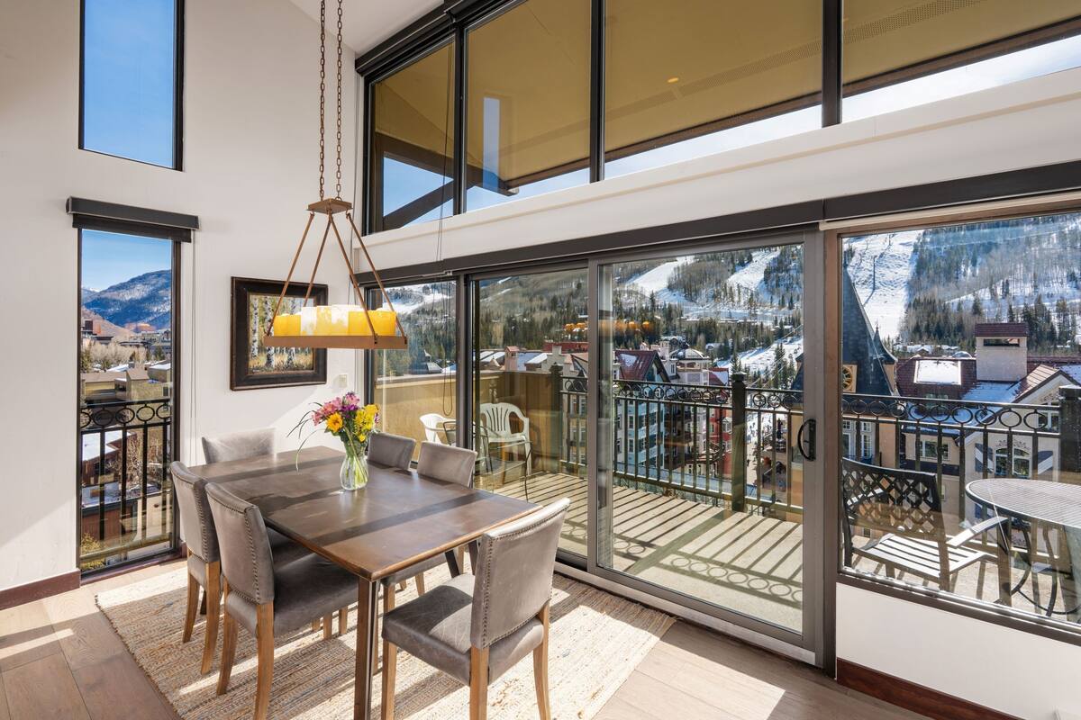A dining area with a table and chairs, a vase with flowers, and a view of a snowy mountain landscape through large windows.