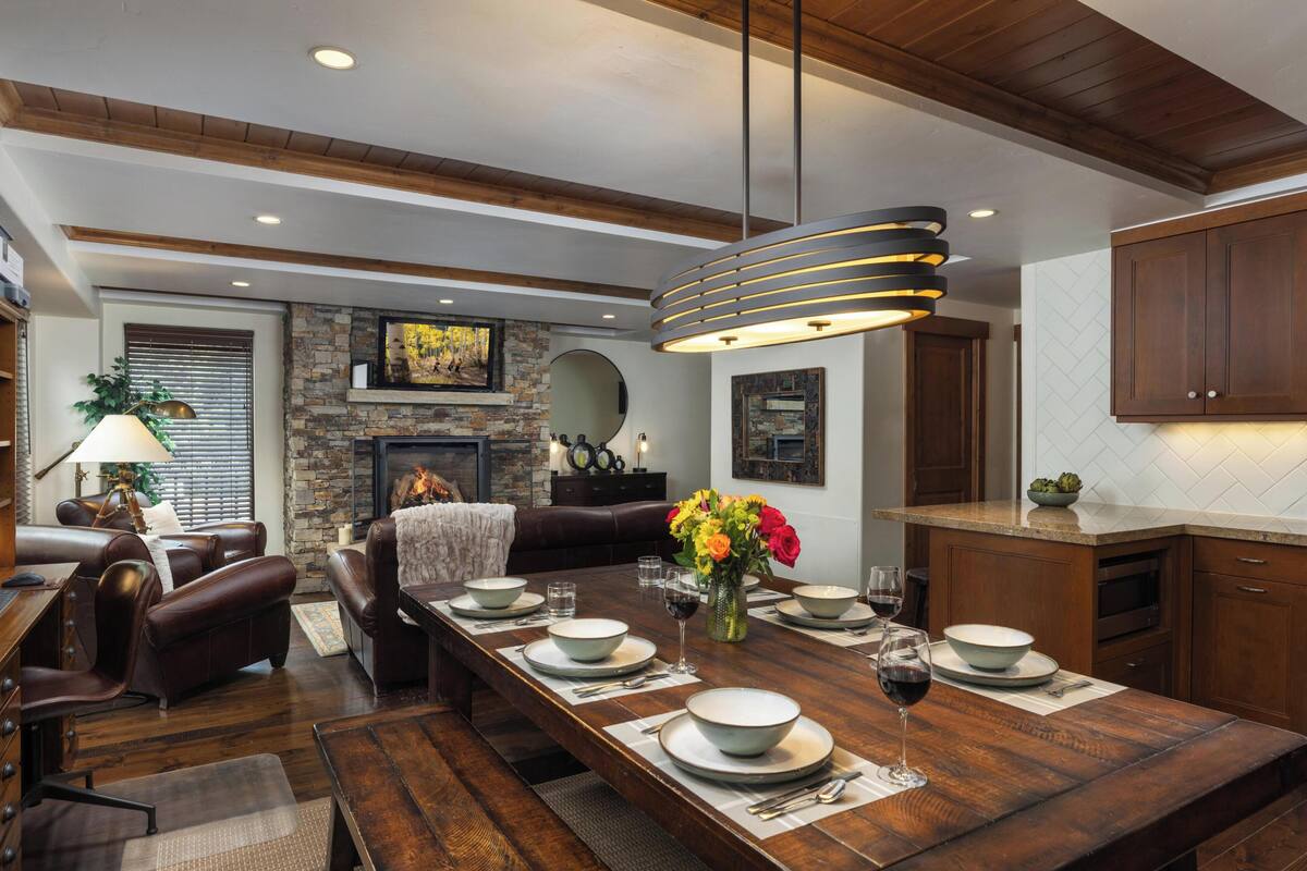 A dining area with a long wooden table set for six, surrounded by leather sofas and a fireplace.