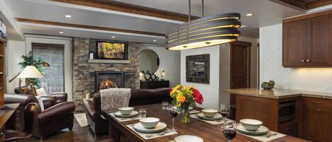 A dining area with a long wooden table set for six, surrounded by leather sofas and a fireplace.