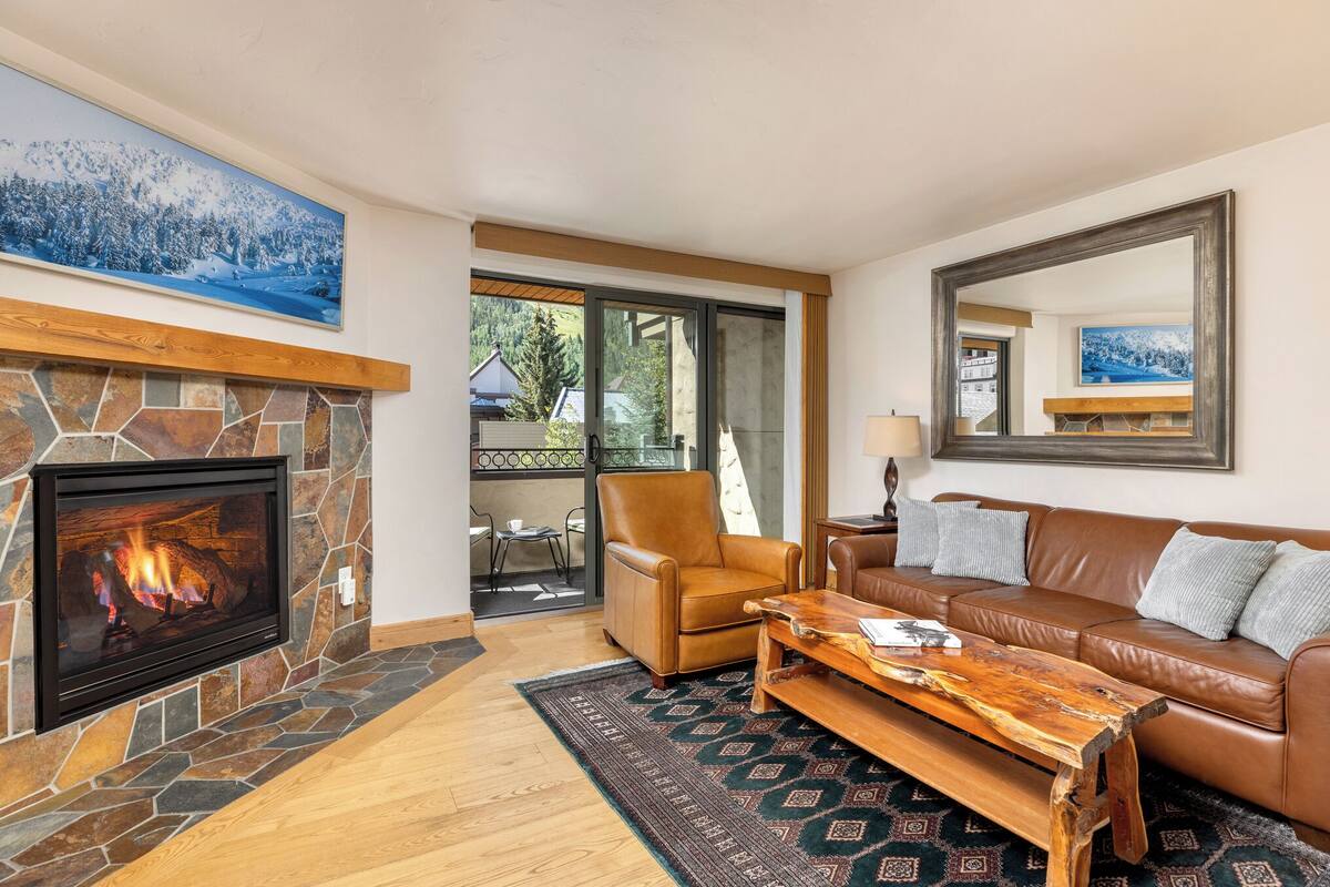 A cozy living room with a stone fireplace, leather sofa, and a large window overlooking a scenic view.