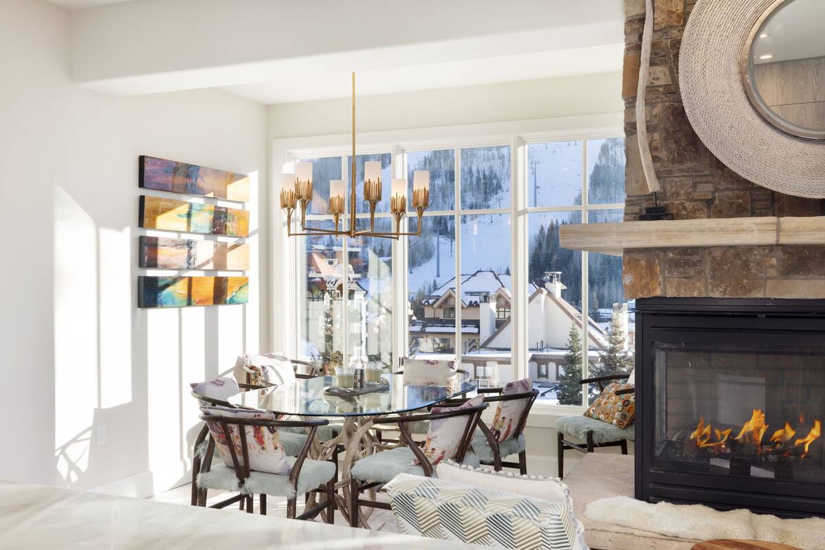A modern living room with a fireplace, a glass dining table, and a view of a snowy landscape through large windows.