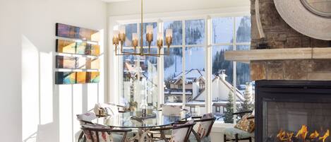 Un salon moderne avec une cheminée, une table à manger en verre et une vue sur un paysage enneigé, visible à travers de grandes fenêtres.