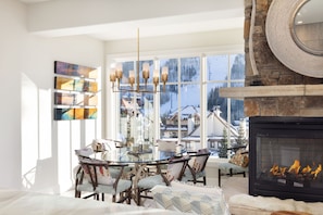 Una habitación luminosa con chimenea, una mesa de comedor con sillas y un ventanal con vistas a casas y árboles.