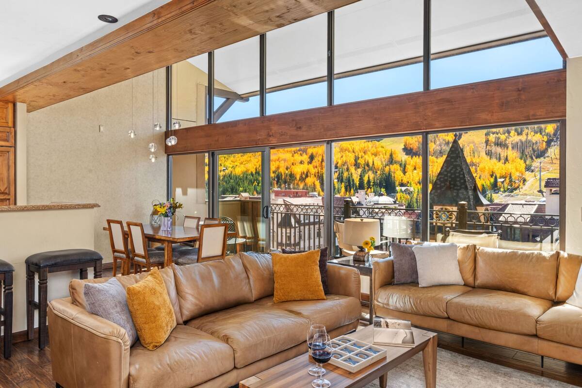 A spacious living room with leather sofas, a wooden coffee table, and large windows offering a view of autumn foliage.