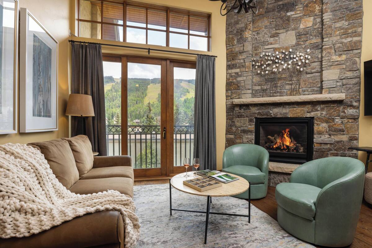 A cozy living room with a stone fireplace, a green leather sofa, a green armchair, and a coffee table with a magazine.