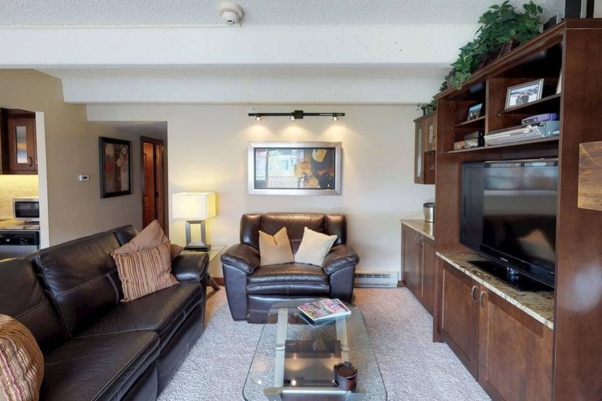 A living room with a leather sofa, a glass coffee table, a television, and a kitchen area with wooden cabinets.