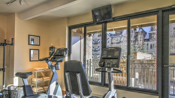 A gym with stationary bikes, a treadmill, and a large window with a view of buildings. - The Landmark (Vail)