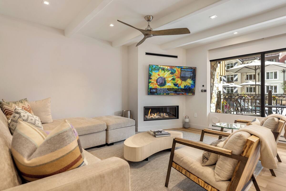 A modern living room with a sofa, armchairs, a coffee table, a fireplace, and a large window with a view of buildings.
