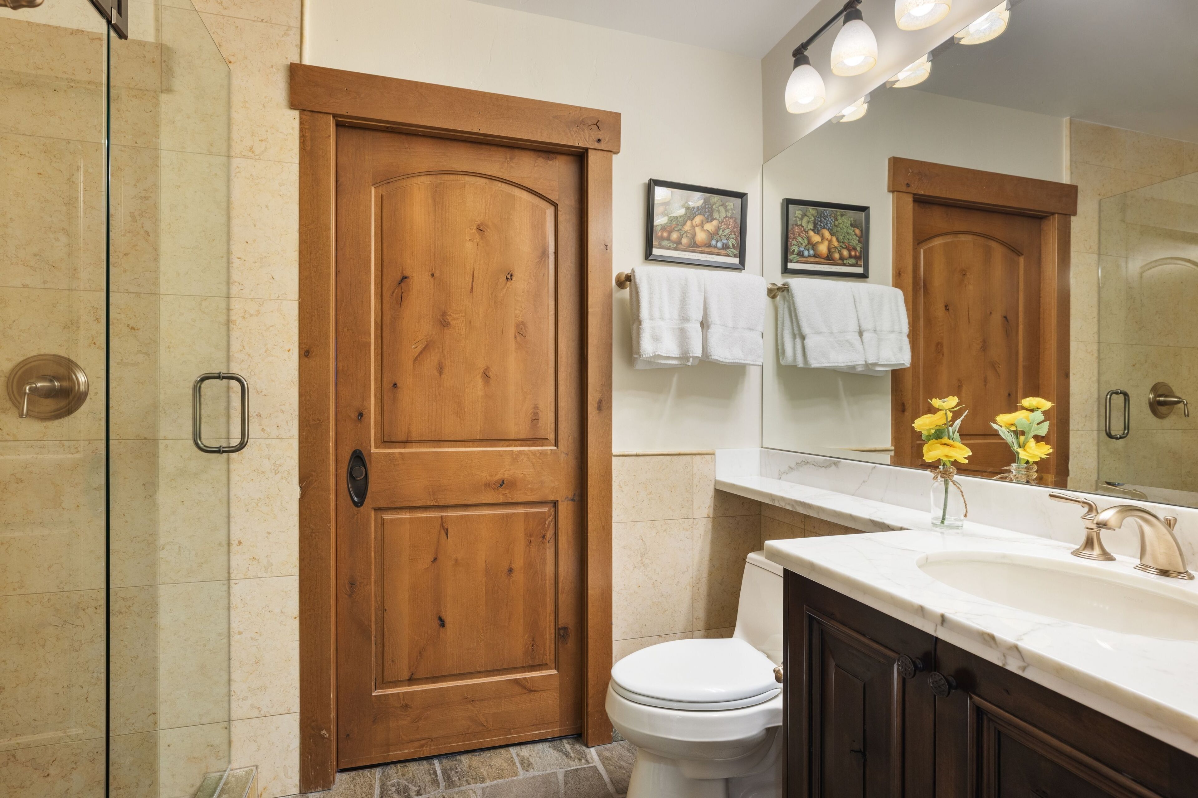 Un baño con puerta de madera, inodoro blanco, lavamanos con espejo y zona de ducha.