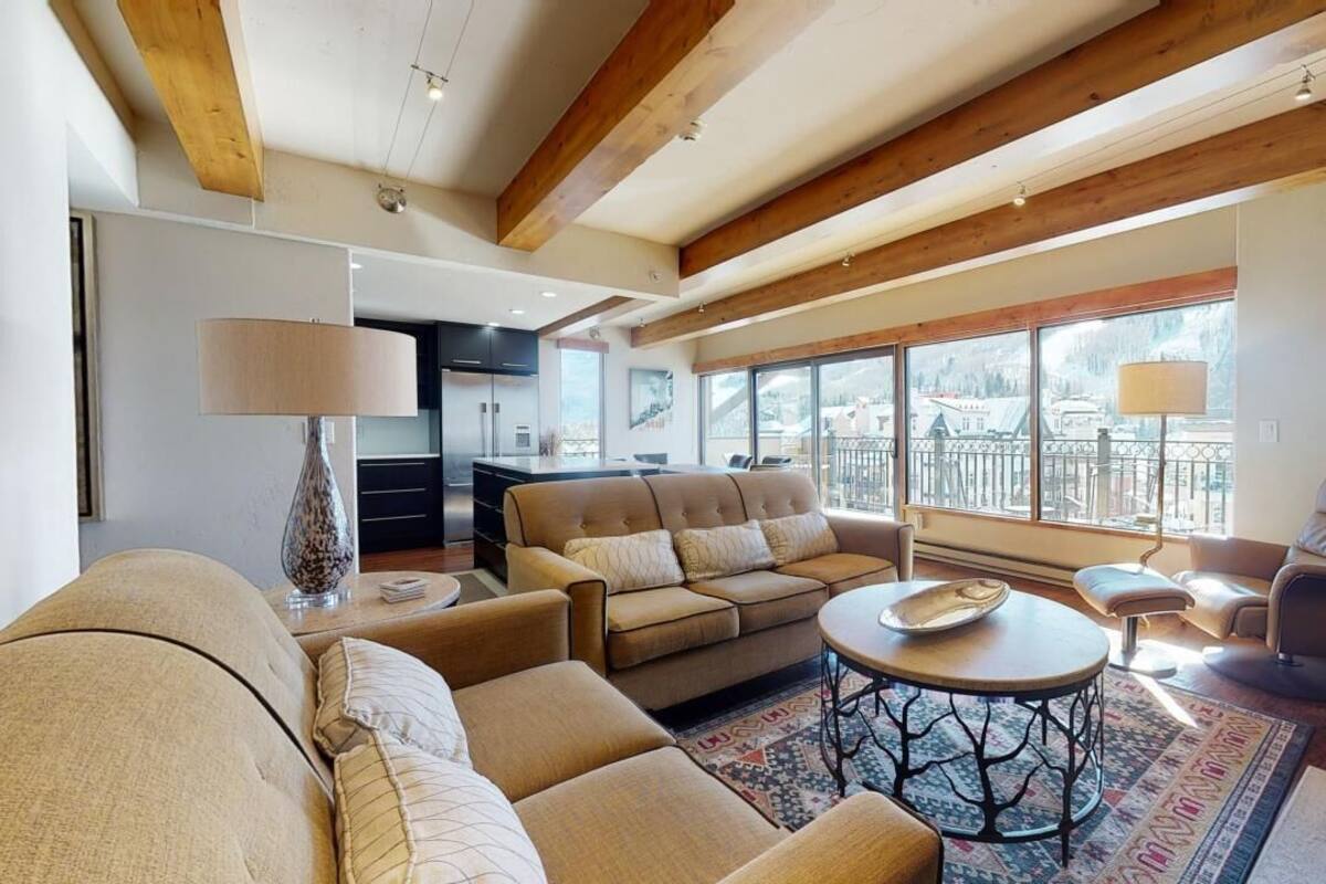 A spacious living room with a sofa, armchairs, and a coffee table. There are large windows offering a view of the mountains.