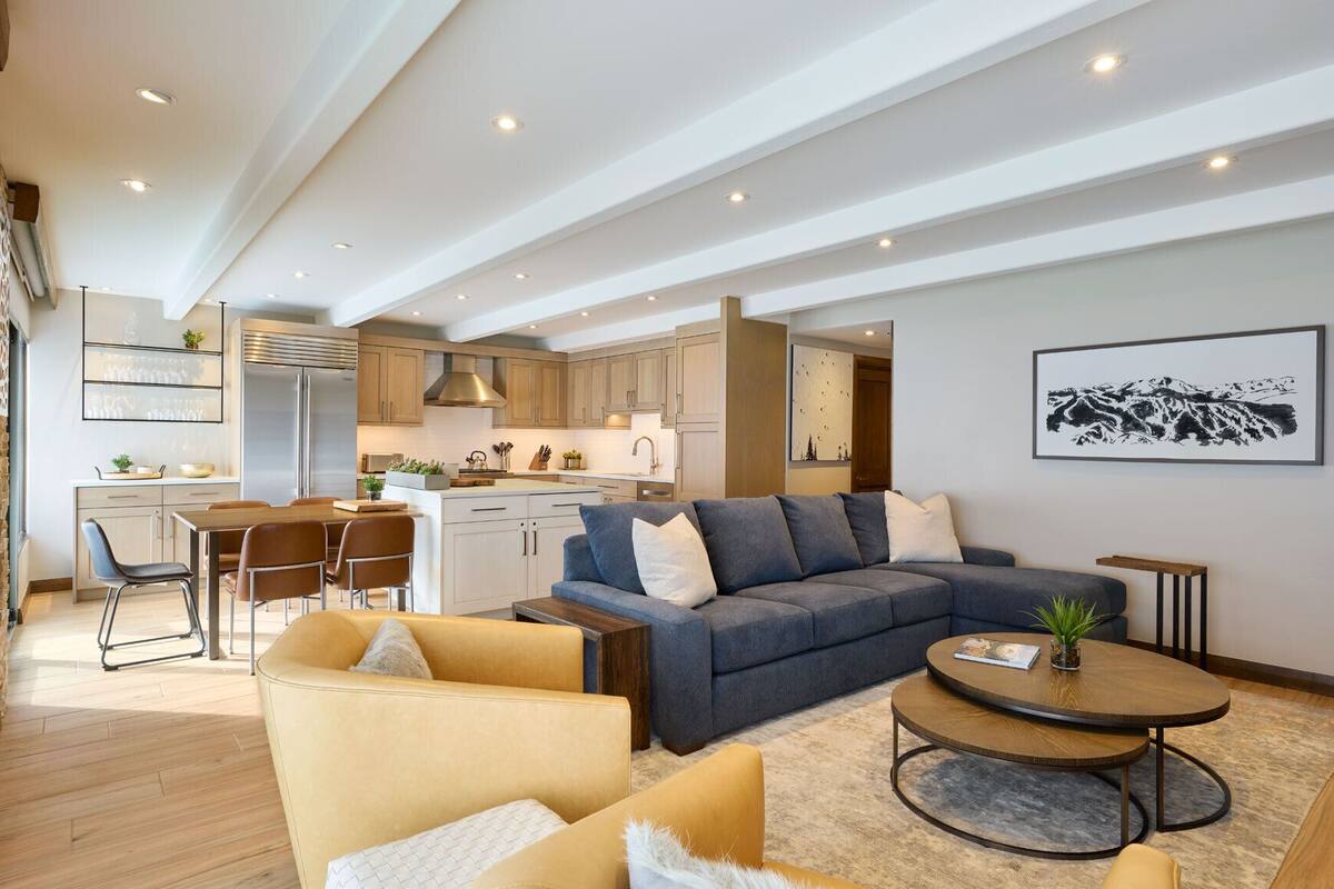 A modern living room with a sofa, coffee table, and chairs. There is a kitchen area with cabinets and a refrigerator in the background.