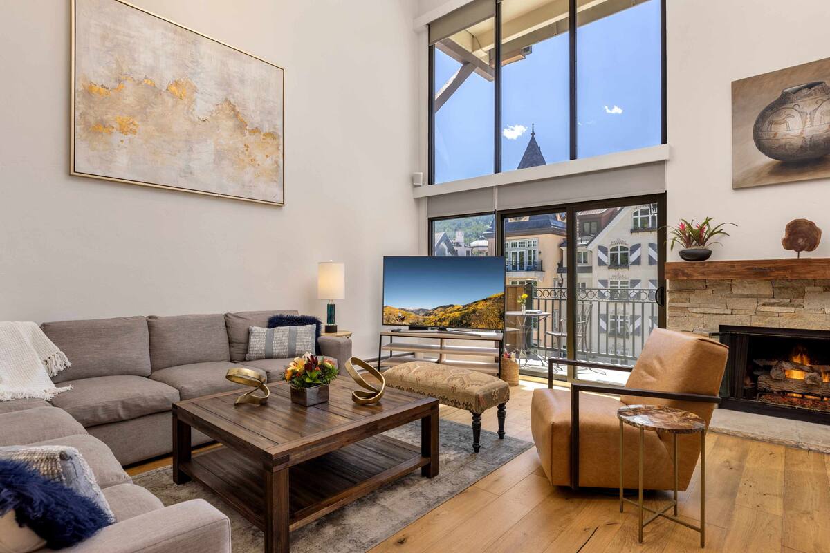A modern living room with a sofa, coffee table, and armchair, a fireplace, and a large window with a city view.