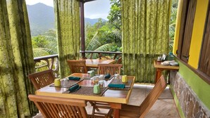 A dining area with wooden tables and chairs, green patterned curtains, and a view of lush greenery and mountains. - Serenity Lodges Dominica (Marigot)