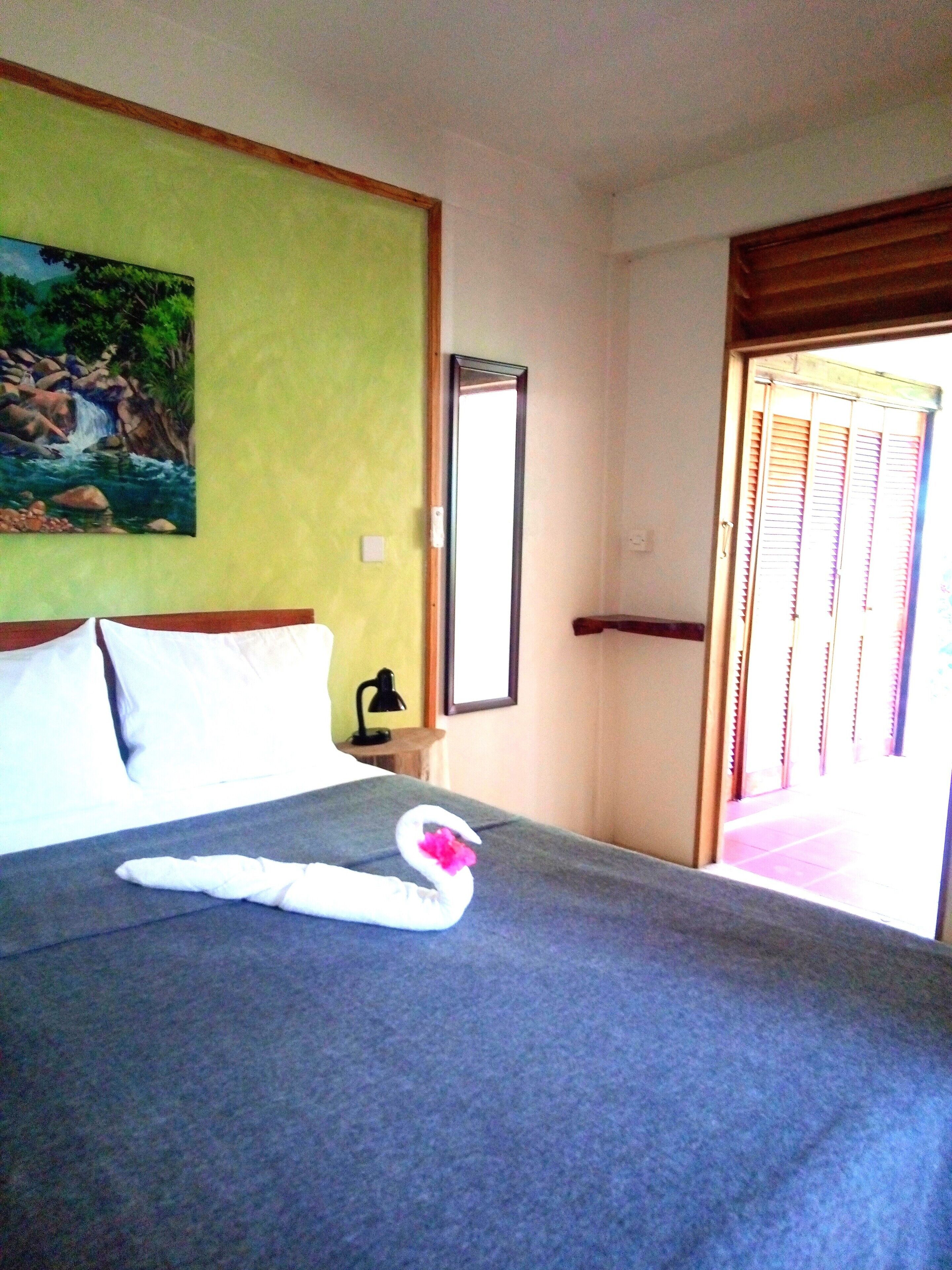 A hotel room with a bed, a painting on the wall, a mirror, and a door with shutters.