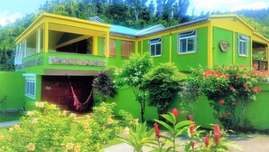 A two-story house with a green exterior and yellow accents, surrounded by lush gardens and trees. - Serenity Lodges Dominica (Marigot)