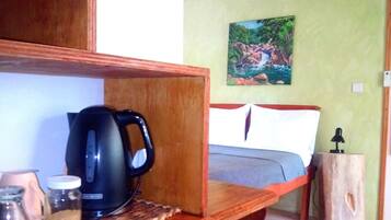 A bedroom with a bed, a nightstand, a painting, a ceiling fan, and a kitchen area with a kettle and jars.