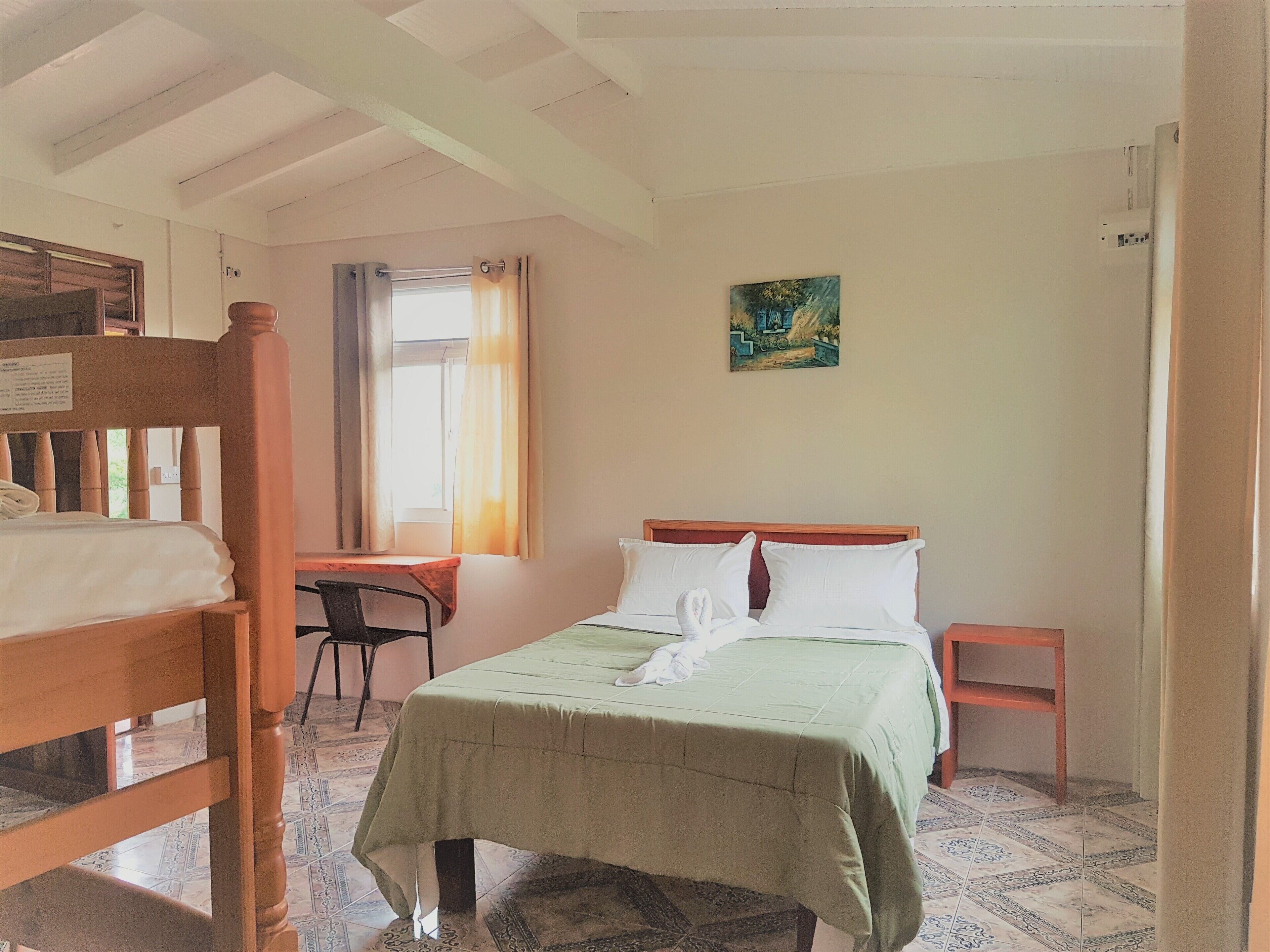 A room with a bed, a desk, and a chair. There is a window with curtains, a painting on the wall, and a tiled floor.