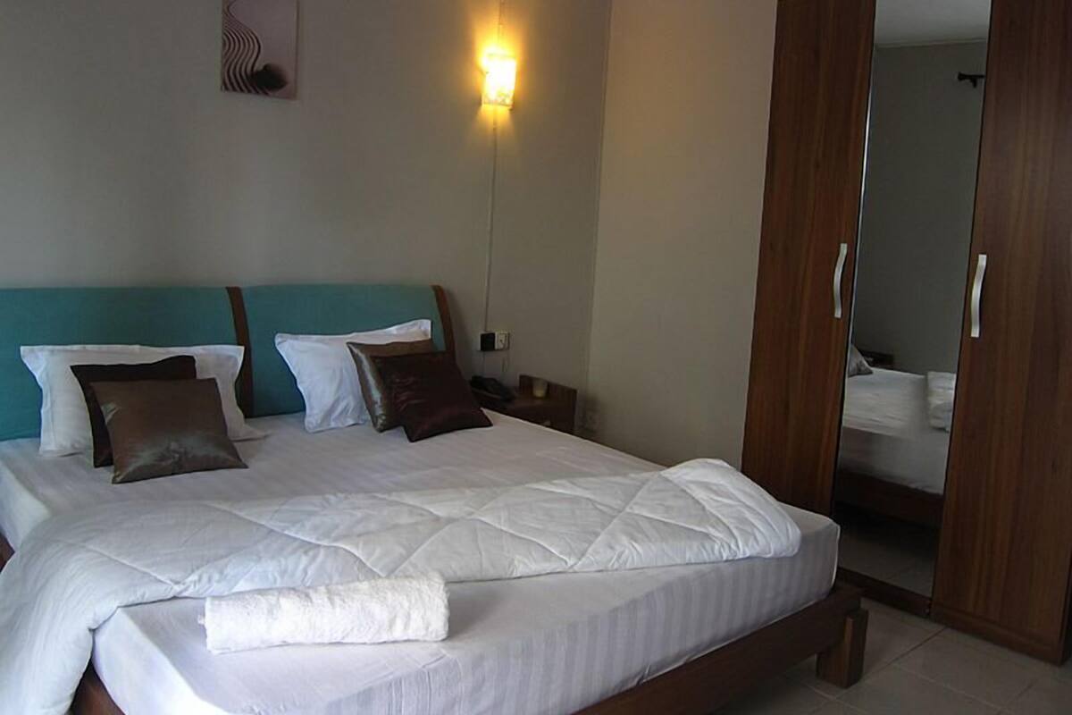 A neatly made bed with white bedding and multiple pillows, a teal headboard, a bedside lamp, and a wooden wardrobe with a mirror.