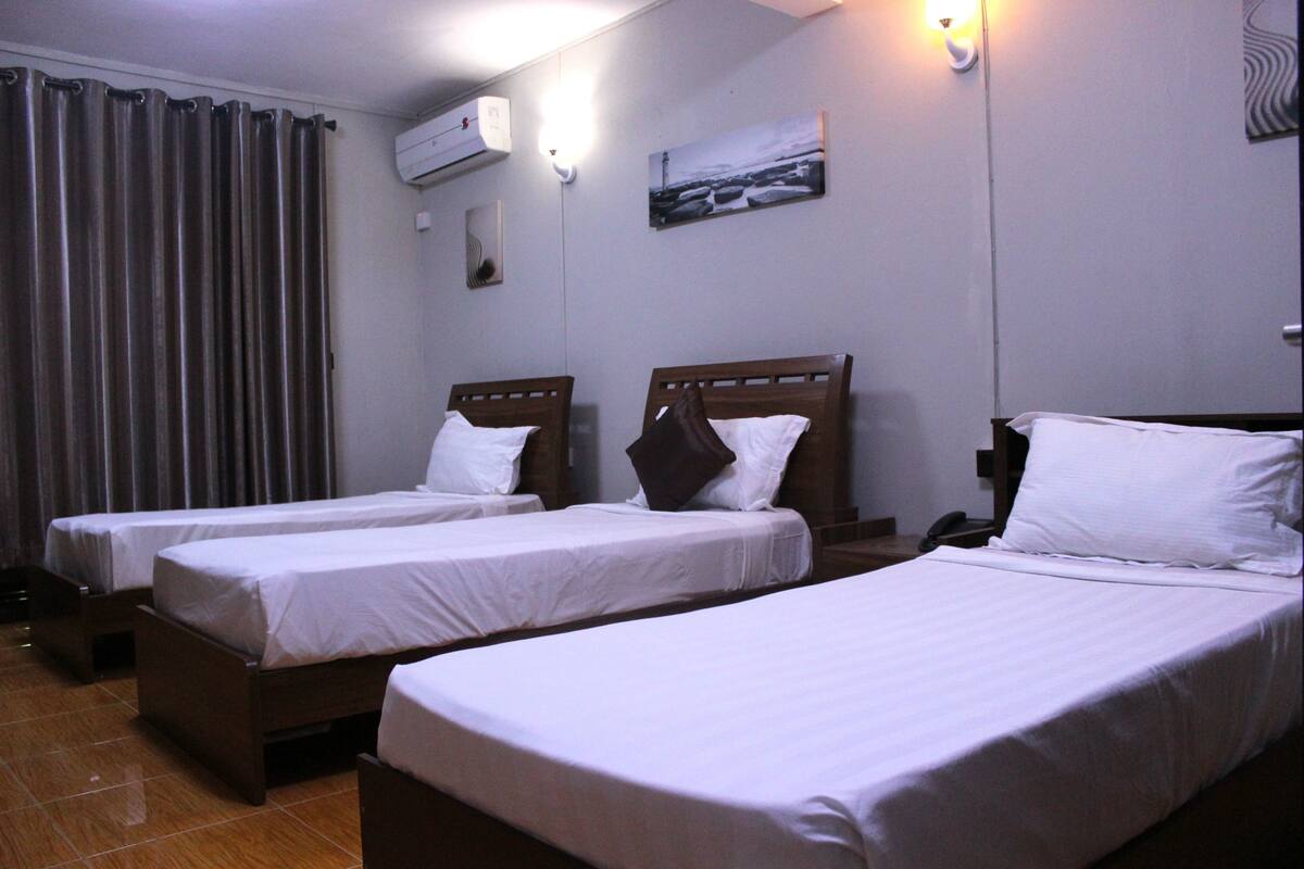 A hotel room with three single beds, a curtain, an air conditioning unit, a painting on the wall, and a telephone on the nightstand.