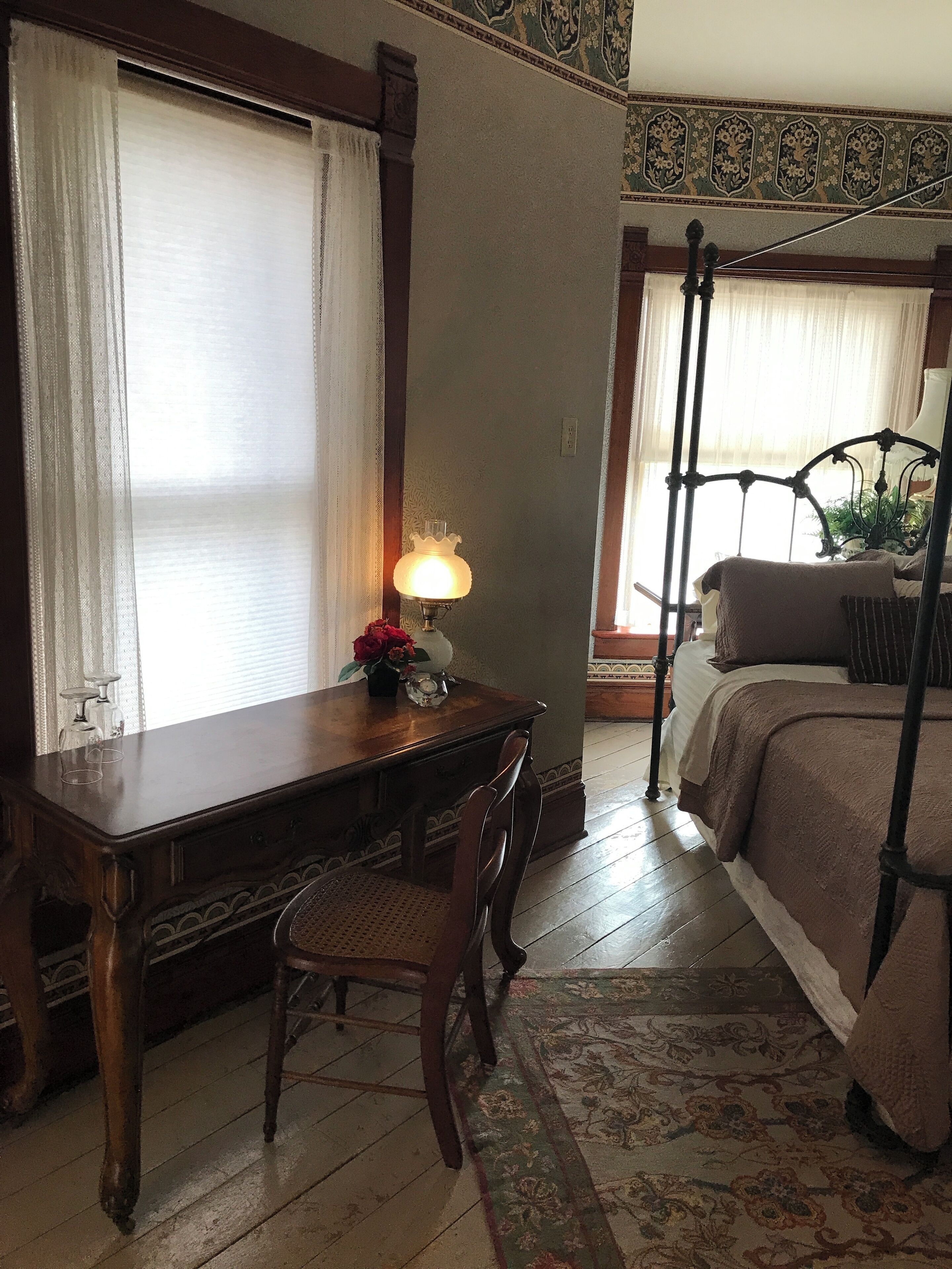 A room with a wooden desk, a chair, a bed with a metal frame, and a window with curtains.