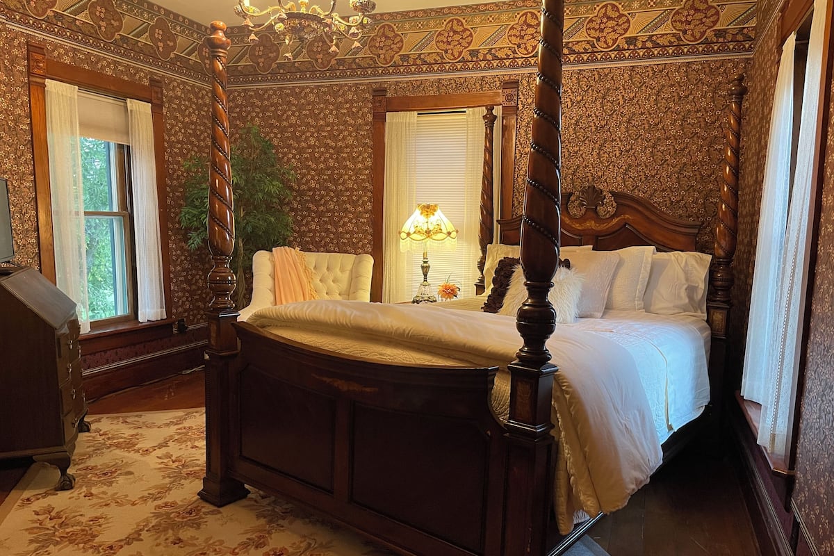 Ein Bett mit Baldachin und einem gepolsterten Kopfteil, eine Holzkommode und ein Teppich mit Blumenmuster.