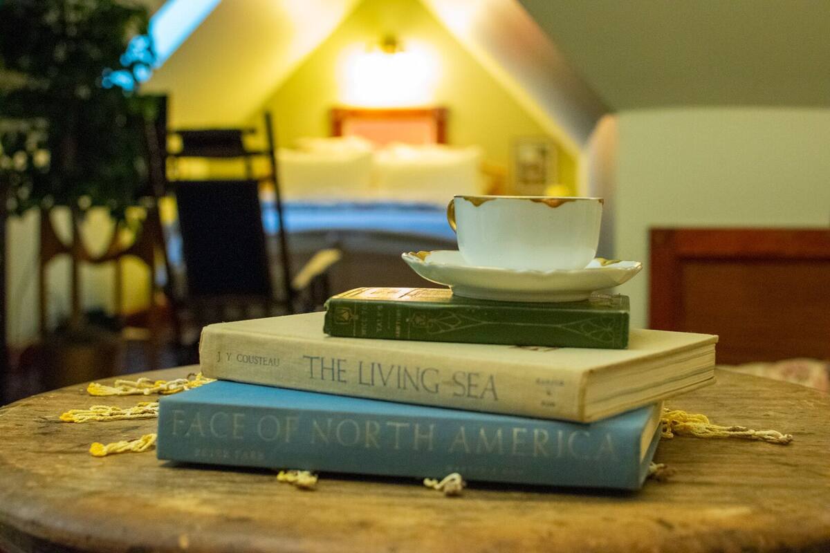 Ein Stapel Bücher auf einem runden Tisch, mit einer Tasse darauf, in einem Zimmer mit Bett und Lampe im Hintergrund.