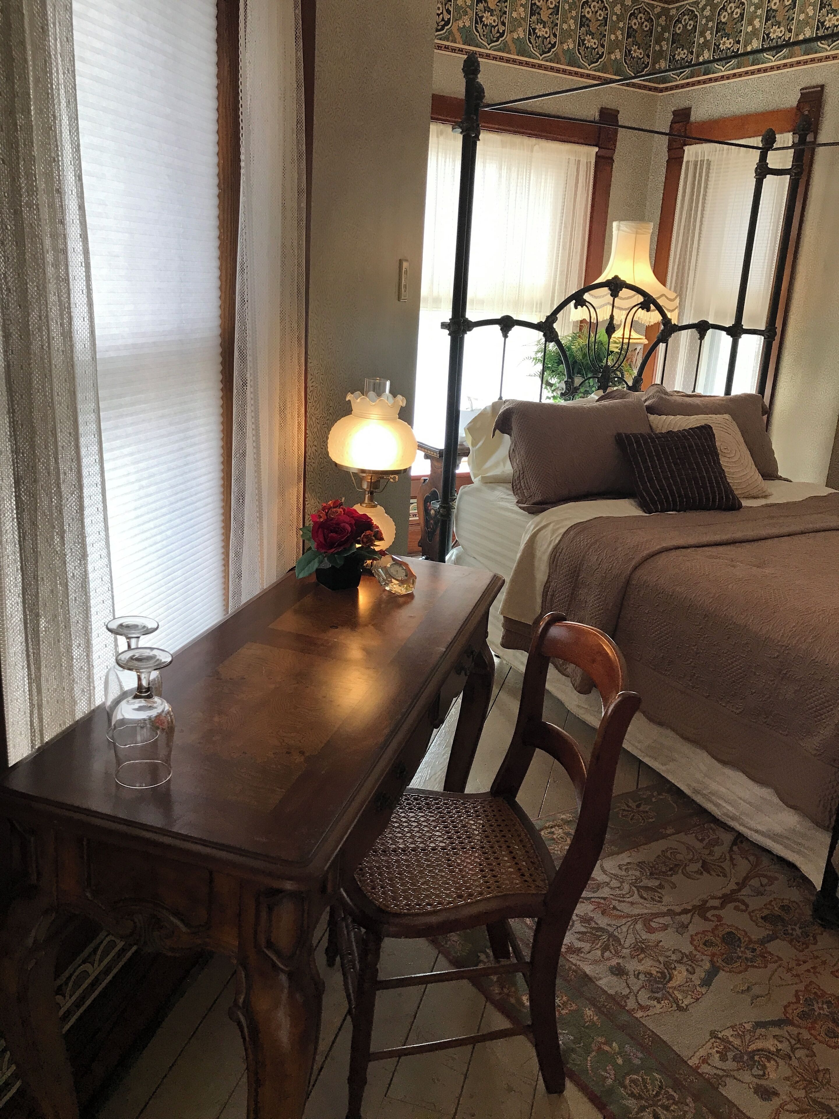 A bedroom with a four-poster bed, a wooden nightstand with a lamp, a chair, and a table with a wine glass.